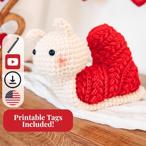 May include: A handmade crochet snail toy with a cream-colored body and a bright red shell. The snail has small black eyes and pink cheeks. The image includes a banner that says "Printable Tags Included!"