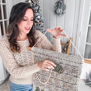 May include: A handmade, gray crochet advent calendar with wooden ornaments and a wooden dowel. The calendar has 24 pockets, each with a wooden ornament numbered 1-24. Green felt Christmas trees are also attached. A string is used for hanging.