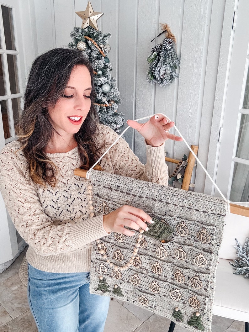 May include: A handmade, gray crochet advent calendar with wooden ornaments and a wooden dowel. The calendar has 24 pockets, each with a wooden ornament numbered 1-24. Green felt Christmas trees are also attached. A string is used for hanging.