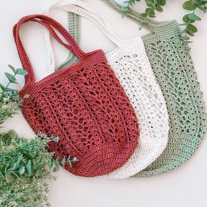 Puede incluir: Tres bolsas de mano de ganchillo en tonos rojo, blanco y verde. Las bolsas tienen patrones de tejido abierto y asas largas. Las bolsas se muestran sobre una superficie blanca con follaje verde.