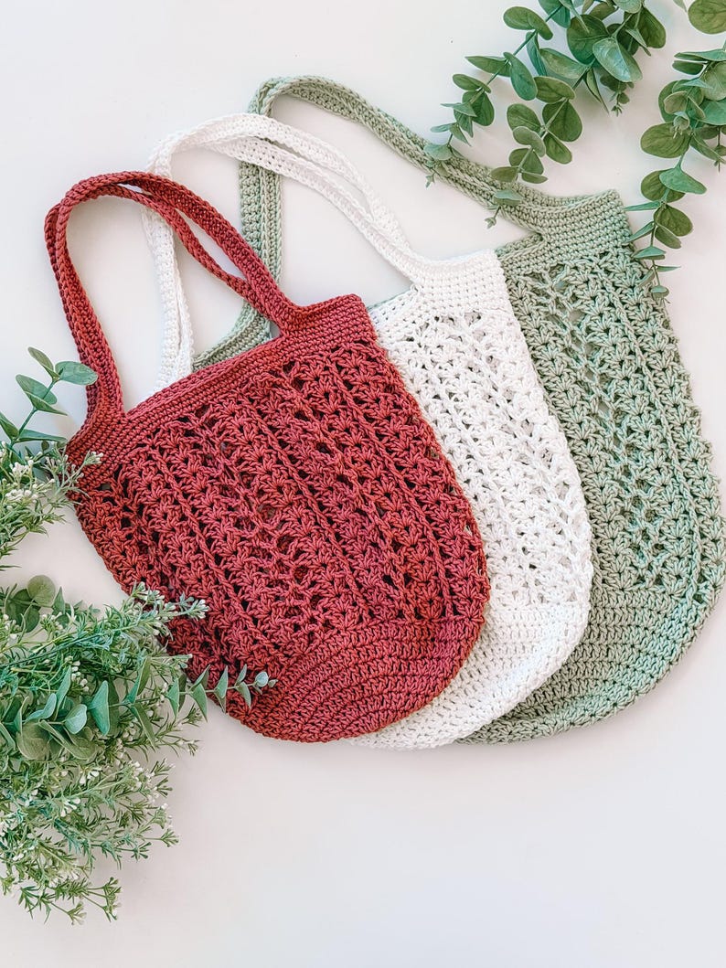 Puede incluir: Tres bolsas de mano de ganchillo en tonos rojo, blanco y verde. Las bolsas tienen patrones de tejido abierto y asas largas. Las bolsas se muestran sobre una superficie blanca con follaje verde.
