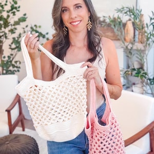 A woman stands indoors holding two crocheted tote bags, one white and one pink—perfect as a handmade gift—amidst plants and chairs in the background.