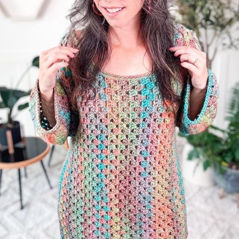 A person is wearing a colorful granny stitch crochet dress, standing in a room with geometric wall decor and plants in the background, accessorized with unique jewelry.