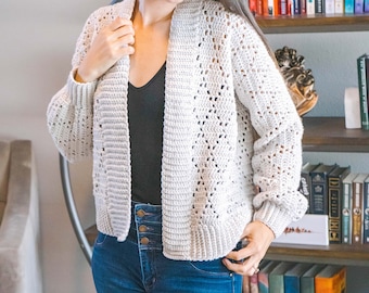 Op de afbeelding: Lichtgrijze gehaakte vest met open voorkant en lange mouwen. Het vest heeft een textuursteekpatroon en wordt gedragen met een zwarte top en een blauwe jeans. De mouwen hebben een lichte pof bij de pols. Het vest is ongeveer 61 cm lang.