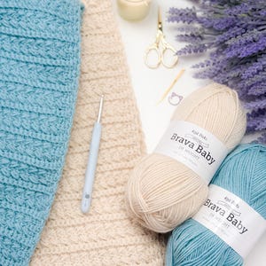 May include: A crafting still life featuring a light blue and cream-colored crochet projects, two balls of "Brava Baby" yarn, a crochet hook, scissors, a candle, and lavender. The yarn is DK weight. The scene is set on a white surface.