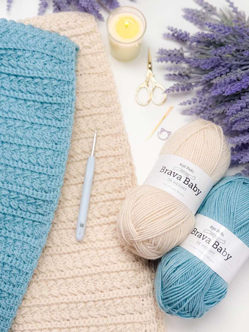 May include: A crafting still life featuring a light blue and cream-colored crochet projects, two balls of "Brava Baby" yarn, a crochet hook, scissors, a candle, and lavender. The yarn is DK weight. The scene is set on a white surface.