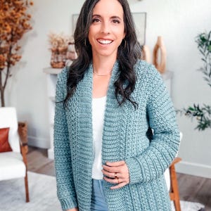 May include: A light blue crochet cardigan with a textured stitch pattern. The long-sleeved cardigan is worn over a white top and blue jeans. The woman in the image is smiling and wearing gold jewelry.