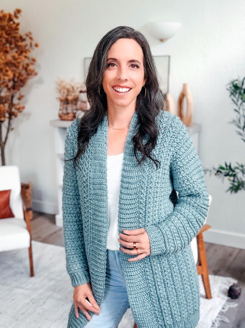 May include: A light blue crochet cardigan with a textured stitch pattern. The long-sleeved cardigan is worn over a white top and blue jeans. The woman in the image is smiling and wearing gold jewelry.