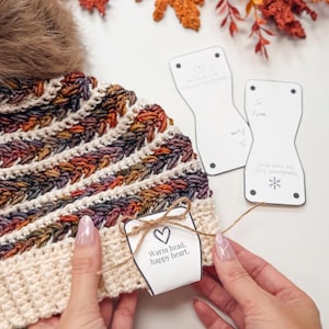 A person ties a tag reading "Warm head, happy heart." onto a knitted hat, with crocheted patterns, autumn leaves, and hints of vintage clothes in the background.