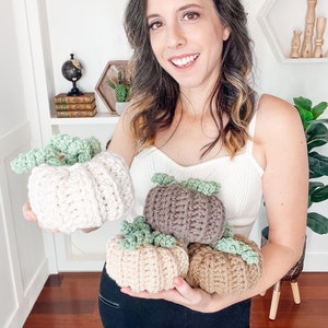 Smiling woman in a white top holding four crochet pumpkins in cream, taupe, beige and tan with green curly stems