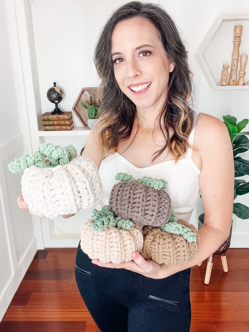 Smiling woman in a white top holding four crochet pumpkins in cream, taupe, beige and tan with green curly stems