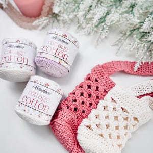 Puede incluir: Tres ovillos de hilo Herrschners cottage cotton en blanco y morado claro, junto a dos bolsas de ganchillo en rosa y crema. Las etiquetas del hilo presentan detalles florales y el texto Herrschners cottage cotton en fuente roja.