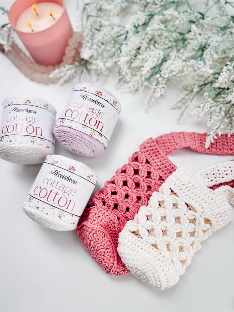 Puede incluir: Tres ovillos de hilo Herrschners cottage cotton en blanco y morado claro, junto a dos bolsas de ganchillo en rosa y crema. Las etiquetas del hilo presentan detalles florales y el texto Herrschners cottage cotton en fuente roja.