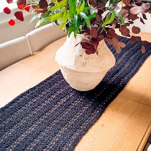 A cream-colored vase with red and green foliage sits on a wooden table with a dark woven table runner, creating the perfect setting for displaying unique jewellery or presenting a thoughtful handmade gift.