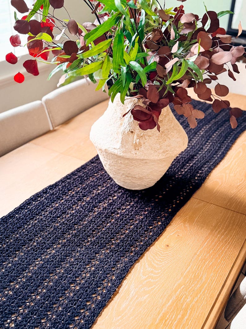 A cream-colored vase with red and green foliage sits on a wooden table with a dark woven table runner, creating the perfect setting for displaying unique jewellery or presenting a thoughtful handmade gift.