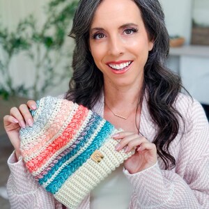 A woman with long, wavy dark hair and a light pink sweater holds up a crochet hat with colorful stripes—a perfect handmade gift. Indoors, with plants and a blurred background.