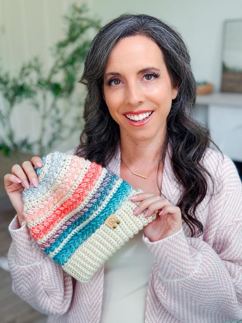A woman with long, wavy dark hair and a light pink sweater holds up a crochet hat with colorful stripes—a perfect handmade gift. Indoors, with plants and a blurred background.