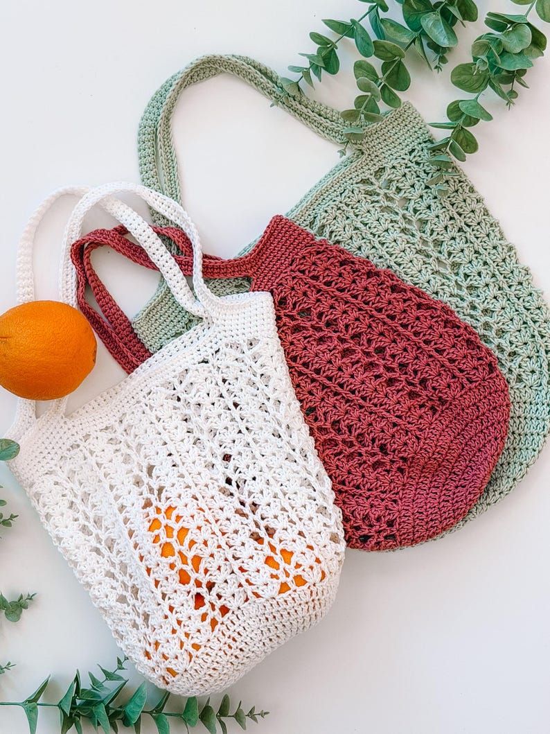 Puede incluir: Tres bolsas de mano de ganchillo en blanco, burdeos y verde salvia. La bolsa blanca contiene naranjas. Las bolsas tienen asas largas y patrones de tejido abierto. Ramas de eucalipto en el fondo. Estas bolsas reutilizables son perfectas para ir de compras.