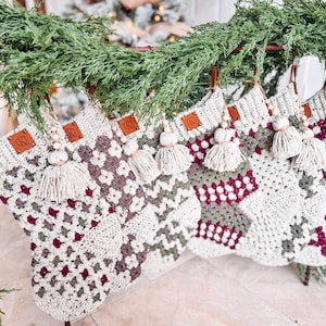 May include: A row of five crocheted Christmas stockings featuring a granny square pattern in cream, sage green, and deep burgundy. Each stocking has a cream-colored cuff, a brown leather tag with the letters KR, and a cream tassel with wooden beads.