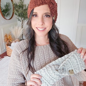 May include: Two knitted headbands, one brown and one gray, with a braided pattern. The brown headband has a button detail. The gray headband has a small leather tag with the letters "KD" on it.