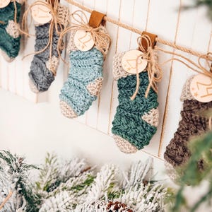 Puede incluir: Una serie de calcetines de Navidad de ganchillo hechos a mano en varios tonos de azul, verde y marrón, colgados de una cuerda. Cada calcetín está adornado con una etiqueta de madera y atado con hilo. Fondo blanco.