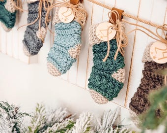 May include: A string of handmade crochet Christmas stockings in various shades of blue, green, and brown, hanging from a rope. Each stocking is adorned with a wooden tag and tied with twine. The stockings are suspended against a white background.