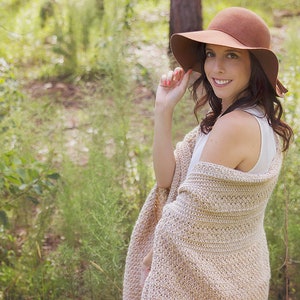 A woman wearing a cream crochet shawl wrap and hat, in a field of greenery.