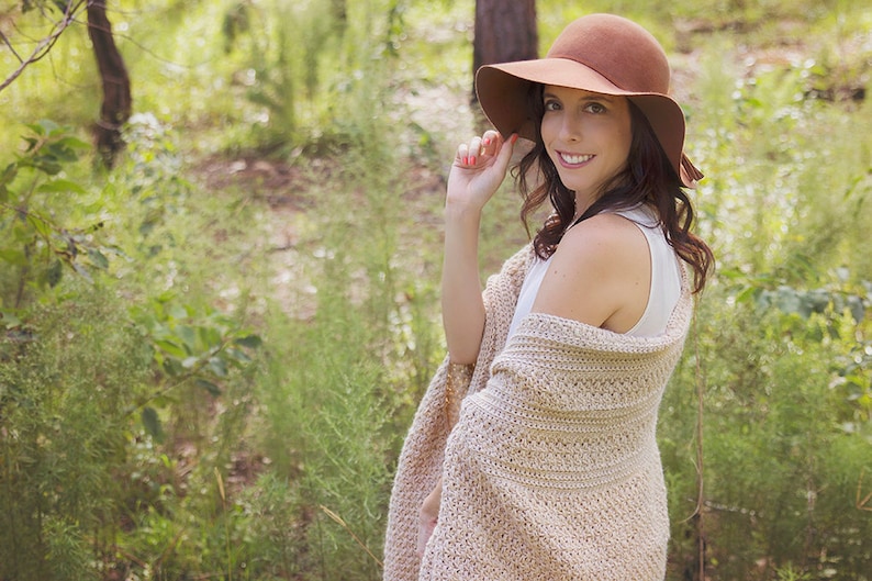 A woman wearing a cream crochet shawl wrap and hat, in a field of greenery.