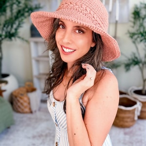 Può includere: Un cappello da sole all'uncinetto color rosa polvere con un dettaglio a fascia intrecciata testurizzata attorno alla corona e una tesa larga e flessibile. Il cappello è realizzato in filato con un motivo a maglia visibile e finitura opaca.