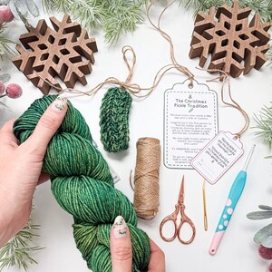 May include: A green yarn skein, a small crocheted green pickle ornament, a spool of twine, copper-coloured scissors, a gold yarn needle, a light blue crochet hook with pink dots, and a tag titled The Christmas Pickle Tradition.