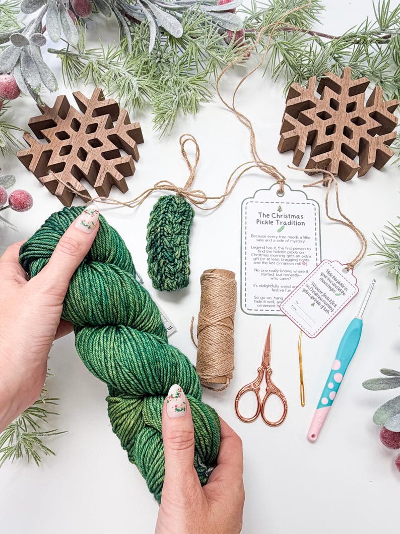 May include: A green yarn skein, a small crocheted green pickle ornament, a spool of twine, copper-coloured scissors, a gold yarn needle, a light blue crochet hook with pink dots, and a tag titled The Christmas Pickle Tradition.