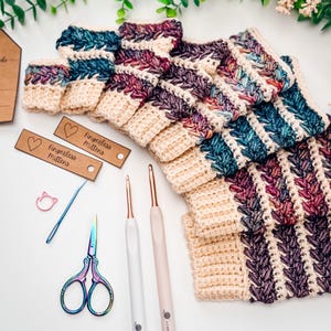 May include: Crocheted fingerless mittens in cream, teal, and purple. The image shows the mittens with crochet hooks, scissors, and tags that read "fingerless mittens". The mittens are made with a textured stitch.
