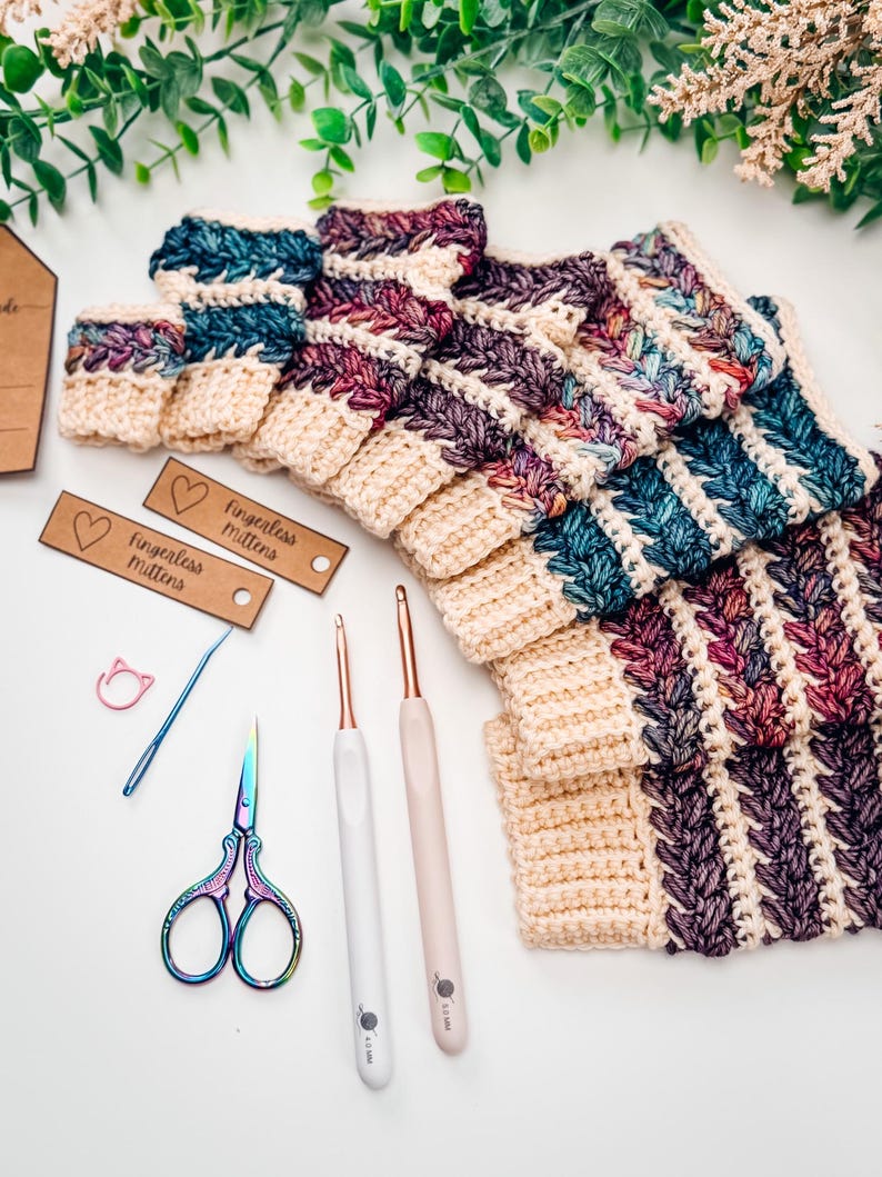 May include: Crocheted fingerless mittens in cream, teal, and purple. The image shows the mittens with crochet hooks, scissors, and tags that read "fingerless mittens". The mittens are made with a textured stitch.
