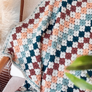 May include: A handmade crochet blanket featuring a classic granny square pattern. The textile is composed of alternating squares in shades of dark teal, muted peach, deep burgundy, and light cream, finished with a simple cream-colored border.