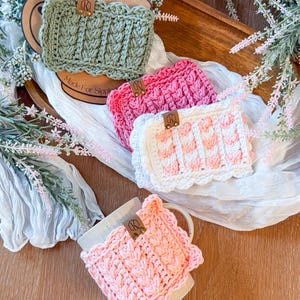 May include: Crocheted mug cozies in shades of green, pink, and white. Each cozy features a textured design and a scalloped edge. A leather tag with the initials "R" is attached to each cozy. The phrase "Made For Sipping" is visible on a cardboard circle.