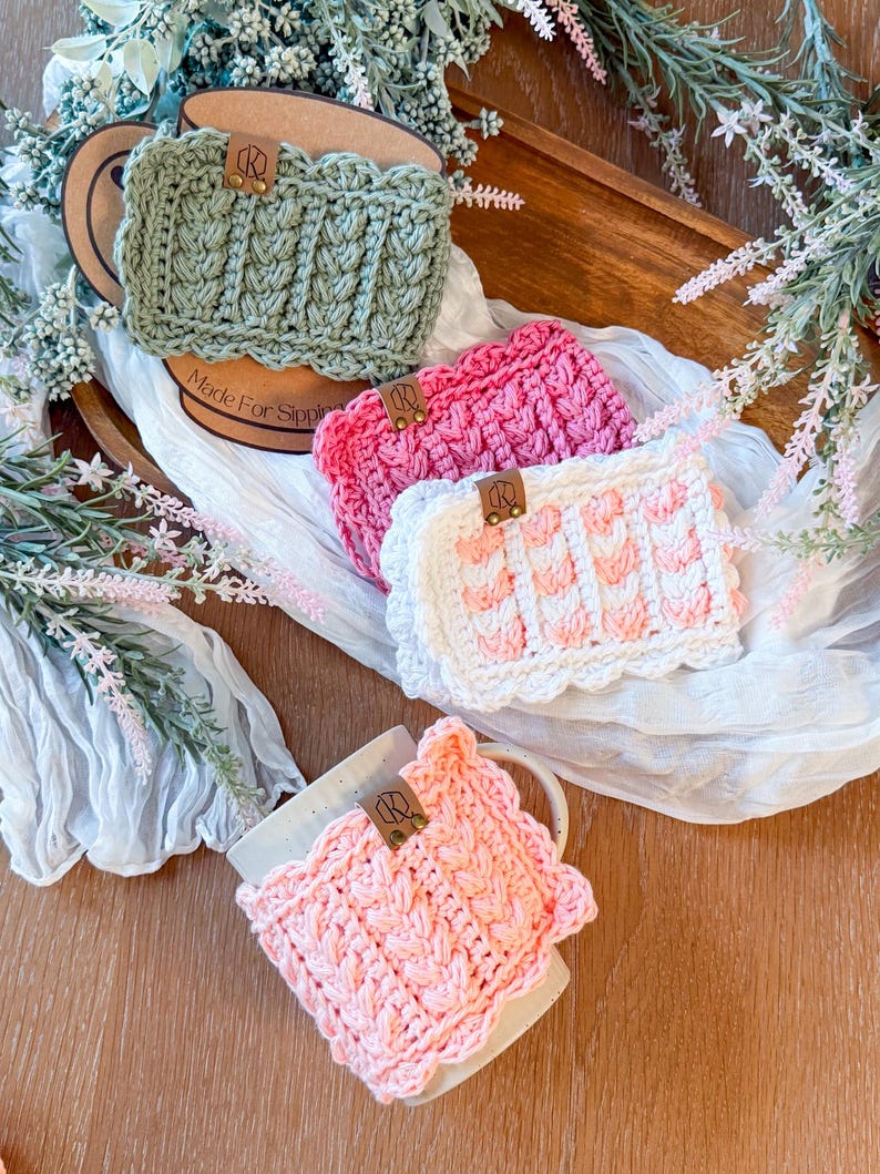 May include: Crocheted mug cozies in shades of green, pink, and white. Each cozy features a textured design and a scalloped edge. A leather tag with the initials "R" is attached to each cozy. The phrase "Made For Sipping" is visible on a cardboard circle.