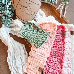 May include: A collection of handmade crochet mug cozies in various colors, including green, peach, red, and white. Each cozy features a textured stitch pattern and a small leather tag. The sign says "Mug Cozy Handmade with Love Made For Sipping."