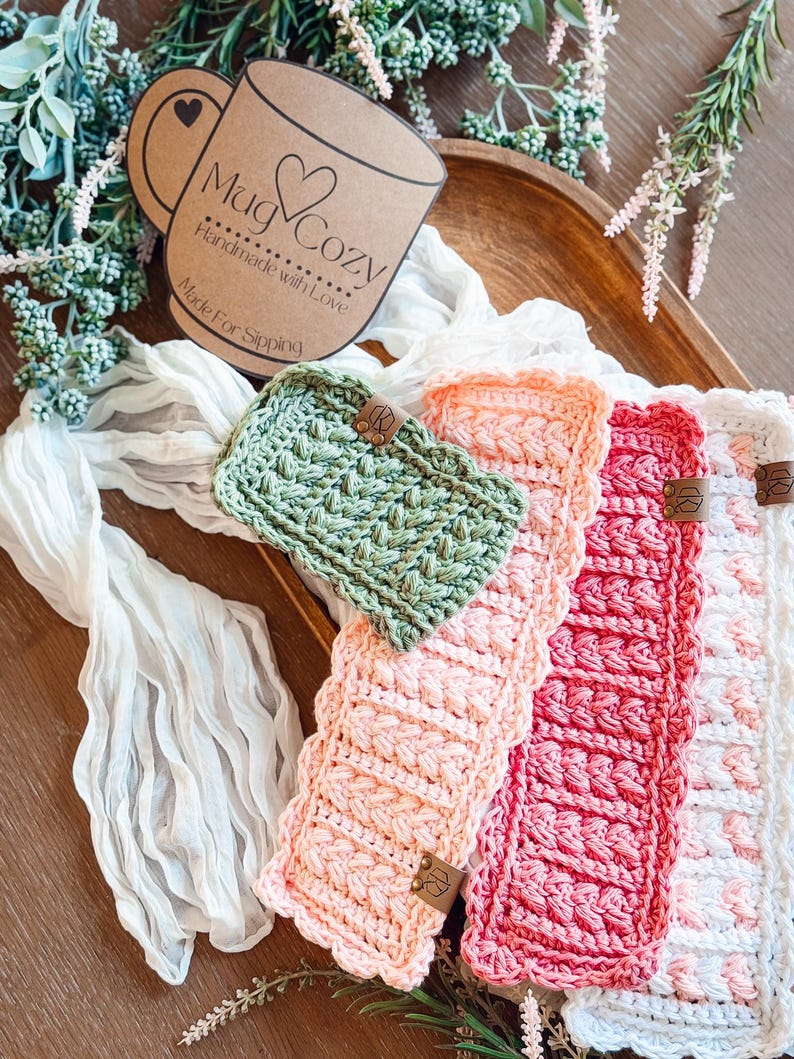 May include: A collection of handmade crochet mug cozies in various colors, including green, peach, red, and white. Each cozy features a textured stitch pattern and a small leather tag. The sign says "Mug Cozy Handmade with Love Made For Sipping."
