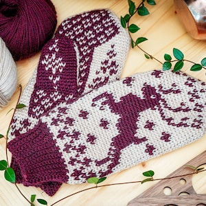 May include: A pair of knitted mittens with a geometric pattern in beige and burgundy. The mittens are lying on a wooden surface with green vines.