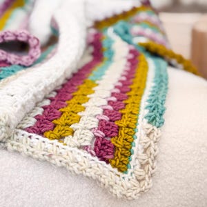 Puede incluir: Una manta de ganchillo multicolor con rayas blancas, azul verdoso, amarillo mostaza y burdeos. La manta está doblada y drapeada sobre una superficie blanca.