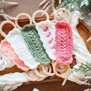 May include: A collection of colorful crochet baby teething rings. The rings are made of natural wood and feature crochet covers in shades of pink, white, and green. The crochet covers have textured patterns and are arranged on a wooden tray.