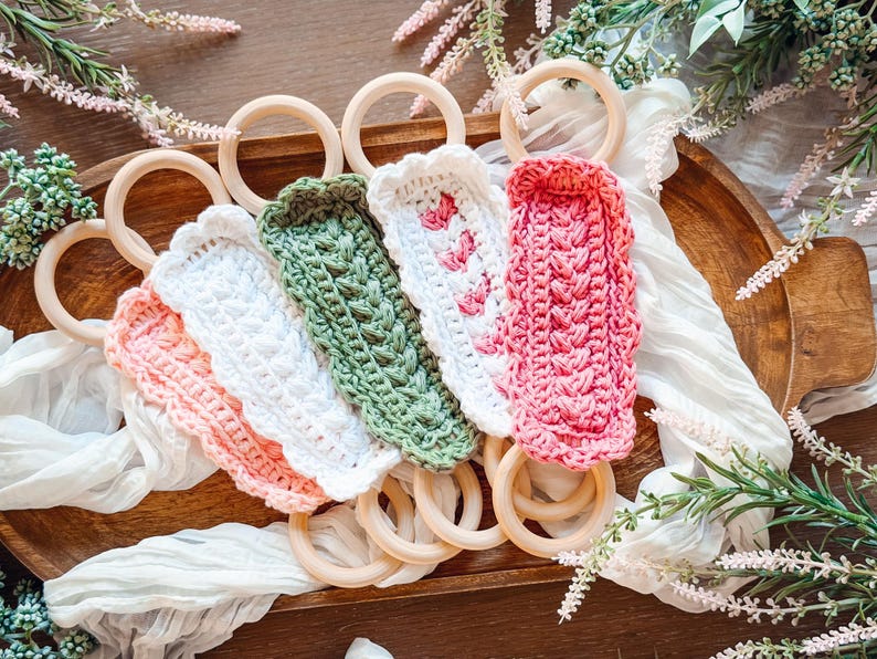 May include: A collection of colorful crochet baby teething rings. The rings are made of natural wood and feature crochet covers in shades of pink, white, and green. The crochet covers have textured patterns and are arranged on a wooden tray.