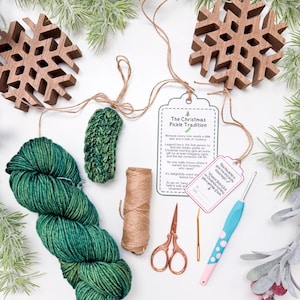 May include: A craft kit featuring a skein of variegated green yarn, a small crocheted green pickle, a spool of brown twine, copper-coloured scissors, a gold needle, and a crochet hook with a blue and pink handle, plus a Christmas Pickle Tradition tag.
