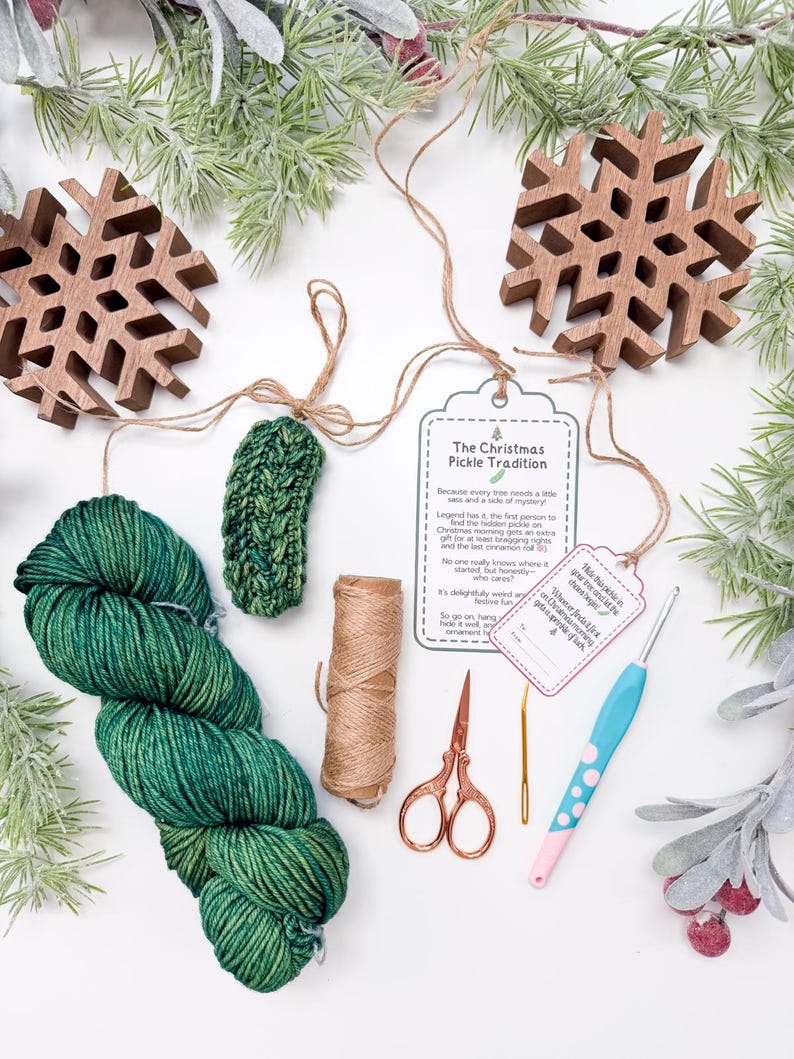 May include: A craft kit featuring a skein of variegated green yarn, a small crocheted green pickle, a spool of brown twine, copper-coloured scissors, a gold needle, and a crochet hook with a blue and pink handle, plus a Christmas Pickle Tradition tag.