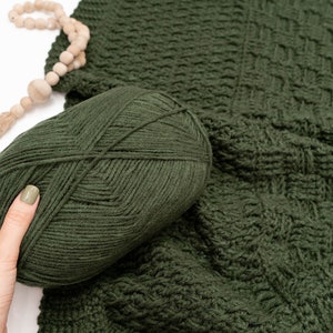 Puede incluir: Un ovillo de lana verde bosque oscuro con aspecto tejido y texturizado, colocado junto a una tela de ganchillo verde oscuro a juego con un patr&oacute;n de rejilla cuadrada y una borla de cuentas de madera marr&oacute;n claro con cuerda de yute.