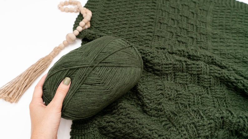 Puede incluir: Un ovillo de lana verde bosque oscuro con aspecto tejido y texturizado, colocado junto a una tela de ganchillo verde oscuro a juego con un patr&oacute;n de rejilla cuadrada y una borla de cuentas de madera marr&oacute;n claro con cuerda de yute.