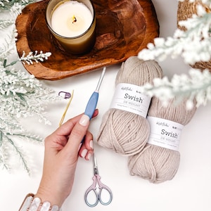 Pode incluir: Uma cena de artesanato aconchegante com uma vela acesa em um copo, uma tigela de madeira e dois novelos de fio bege com a etiqueta "Swish DK Weight". Uma agulha de croch&ecirc;, tesoura e uma pulseira tamb&eacute;m est&atilde;o presentes, sugerindo um projeto de tric&ocirc; ou croch&ecirc;.