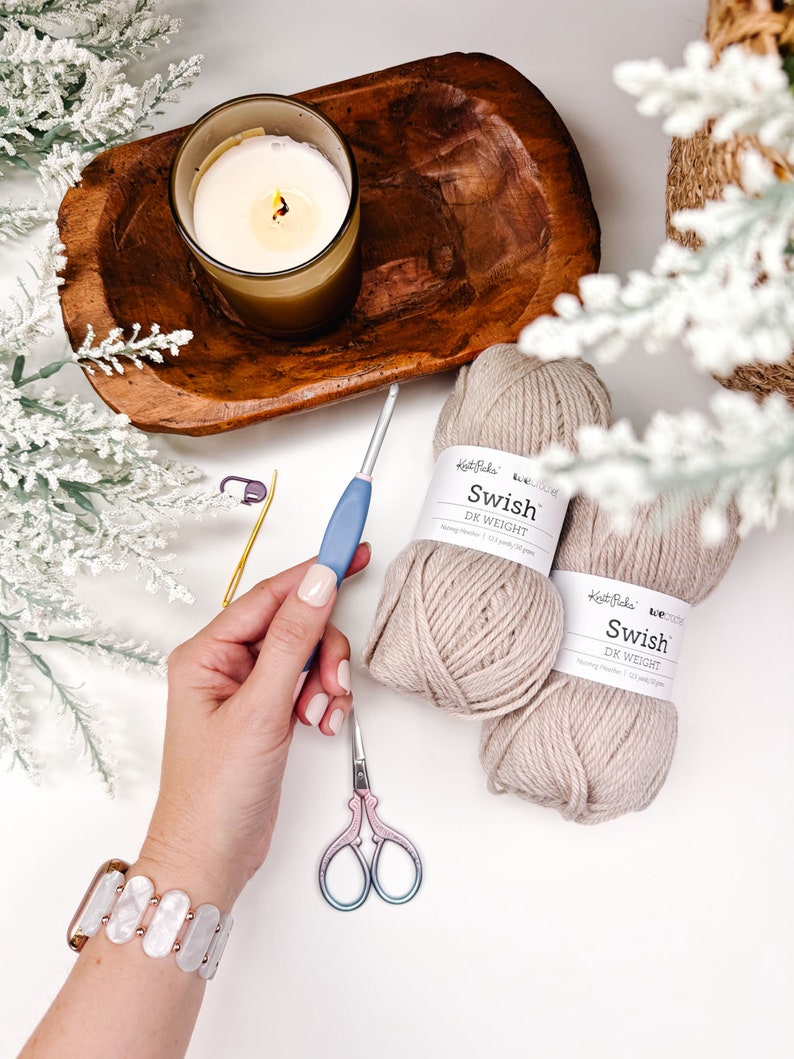 Pode incluir: Uma cena de artesanato aconchegante com uma vela acesa em um copo, uma tigela de madeira e dois novelos de fio bege com a etiqueta "Swish DK Weight". Uma agulha de croch&ecirc;, tesoura e uma pulseira tamb&eacute;m est&atilde;o presentes, sugerindo um projeto de tric&ocirc; ou croch&ecirc;.