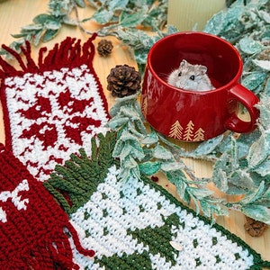 May include: Two crocheted Christmas-themed mug cozies, one red and white with a snowflake design, the other green and white with a Christmas tree design. A small hamster is sitting in a red mug with a Christmas tree design.
