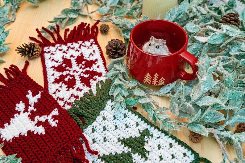 May include: Two crocheted Christmas-themed mug cozies, one red and white with a snowflake design, the other green and white with a Christmas tree design. A small hamster is sitting in a red mug with a Christmas tree design.